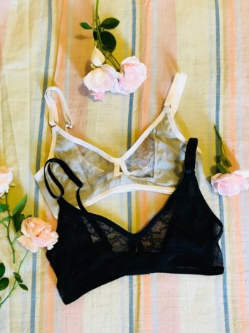 A flat lay photo of two bralettes: the Bully Boy Posey bra in gray and black. The gray bra is made of mesh and lace and features a white piping trim. The black is all black mesh and lace with black trim. Both are adjustable.
