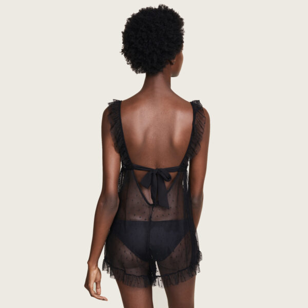 A model (shown back view) wears the black lace Cou Cou Lola tie-back teddy by Only Hearts. It is a sheer polka dot lace teddy with ruffles on the straps and around the bottom of the legs. A princess seam under the bust provides shape and support.