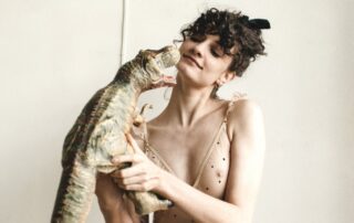 A model wears the Le Petit Trou Clementine Bodysuit and holds a toy Tyrannosaurus Rex. The Clementine bodysuit is made of a sheer light beige covered in tiny black velvet stars and ruffles on the straps.