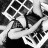 A model poses on a wicker chair wearing the Lena bralette by Hopeless Lingerie. It is a black mesh bra featuring large polka dots and is a classic triangle bralette with a halter strap. She also wears a sheer lacy black skirt with ruffles
