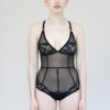 Model standing flat front wearing the Iona bodysuit- a mesh and lace paneled sheer black bodysuit. It also features seams to help the fit be flattering to the body.