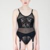 Model facing forward wearing the Rose Garter tank. Rose is a paneled sheer top featuring mesh and lace panels, sdjustable straps and attached garters at the bottom. The model wears beige thigh high tights attached to the garters.