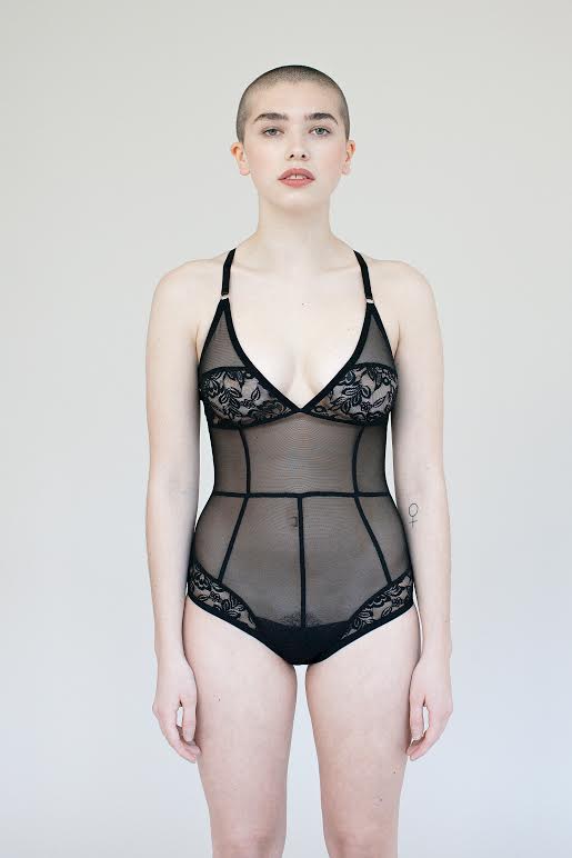 Model standing flat front wearing the Iona bodysuit- a mesh and lace paneled sheer black bodysuit. It also features seams to help the fit be flattering to the body.