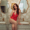 model poses sitting on a counter with ornate mirrors and wallpaper. model wears the kilo brava eyelash teddy in red lace.