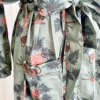 close in view of a mannequin wearing the only hearts luv island kimono robe in moss green with a tropical print. The robe is tied with the belt in a bow and you can see the front pockets as well. The mannequin stands in front of a tropical plant and white doors.