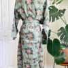 back view of a mannequin wearing the only hearts luv island kimono robe in moss green with a tropical print. The robe is tied with the belt. The mannequin stands in front of a tropical plant and white doors.