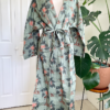 front view of a mannequin wearing the only hearts luv island kimono robe in moss green with a tropical print. The robe is tied with the belt in a bow at the waist. The mannequin stands in front of a tropical plant and white doors.