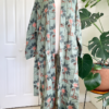 front view of a mannequin wearing the only hearts luv island kimono robe in moss green with a tropical print. The robe is open and untied with the belt hanging loosely at the sides. The mannequin stands in front of a tropical plant and white doors.
