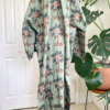 back view of a mannequin wearing the only hearts luv island kaftan in moss green with a tropical print. The mannequin stands in front of a tropical plant and white doors.