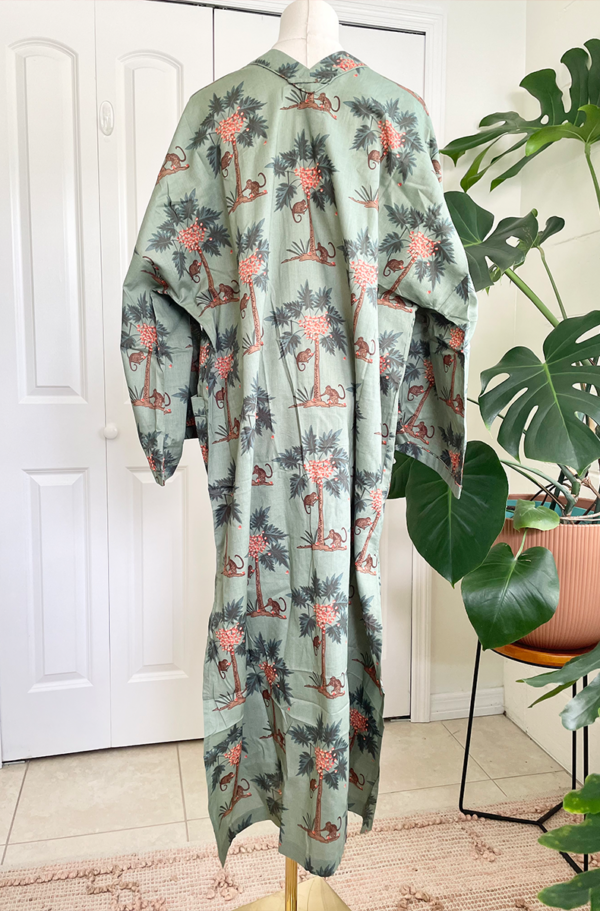 back view of a mannequin wearing the only hearts luv island kaftan in moss green with a tropical print. The mannequin stands in front of a tropical plant and white doors.